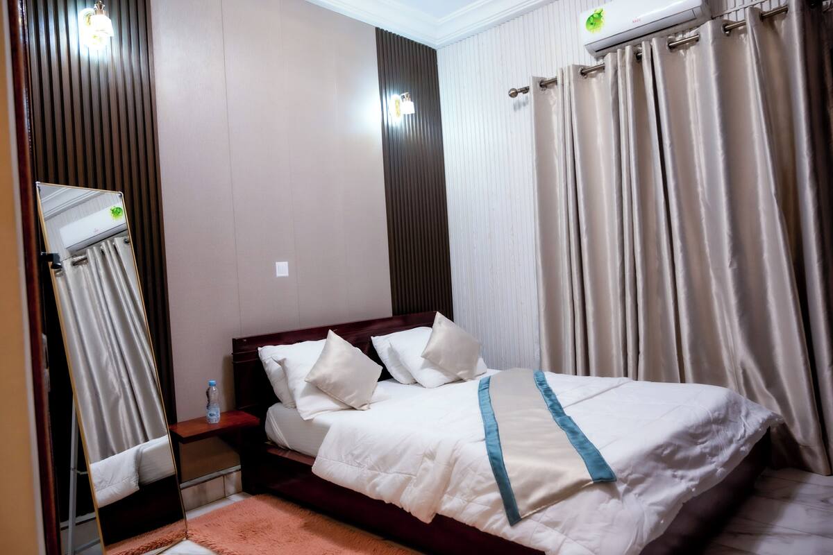 A hotel room with a bed, a bedside table, a mirror, and a window with curtains.
