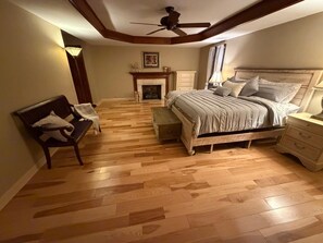 Un dormitorio con piso de madera, una cama con cabecera, un banco, una mesita de noche, un aparador, un ventilador de techo y una chimenea.