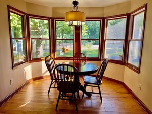 Un comedor con una mesa redonda, cuatro sillas, una lámpara colgante y grandes ventanas con vistas a zonas verdes.