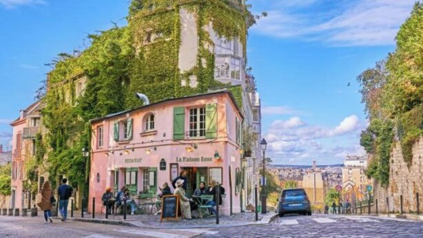 En byggnad med ett café som heter ”La Maison Rose”, med en rosa fasad och gröna fönsterluckor, omgiven av kullerstensgator och grönska.
