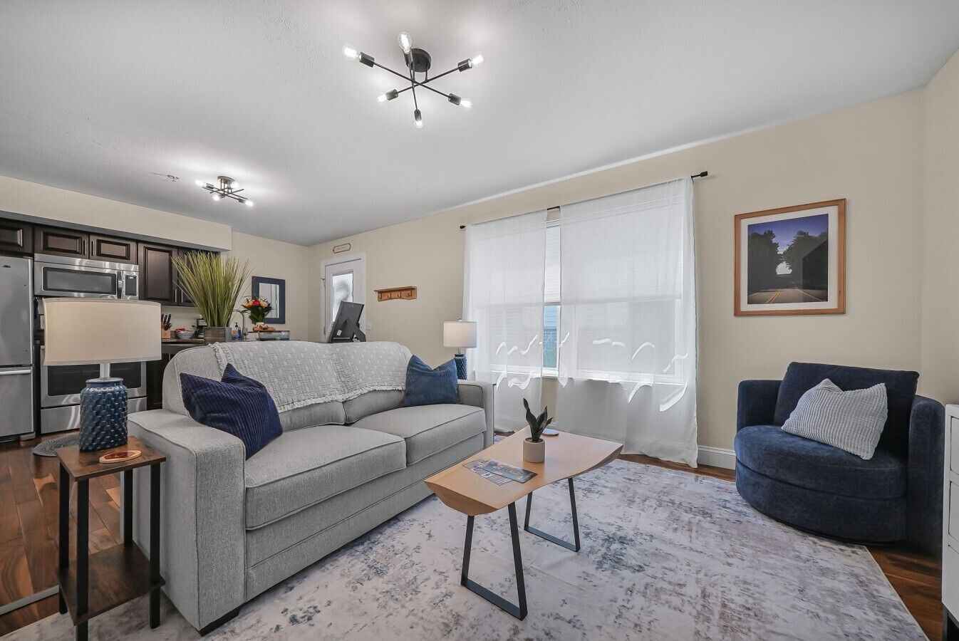 A living room with a grey sofa, a blue armchair, a wooden coffee table, and a kitchen in the background.