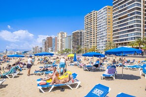 A beach with sun loungers and umbrellas, a clear sky, and high-rise buildings in the background. - Las Rocas 14-A Apartment Levante Beach (Benidorm)