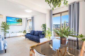 A modern living room with a large TV, a blue sofa, a glass dining table, and potted plants. - Las Rocas 14-A Apartment Levante Beach (Benidorm)