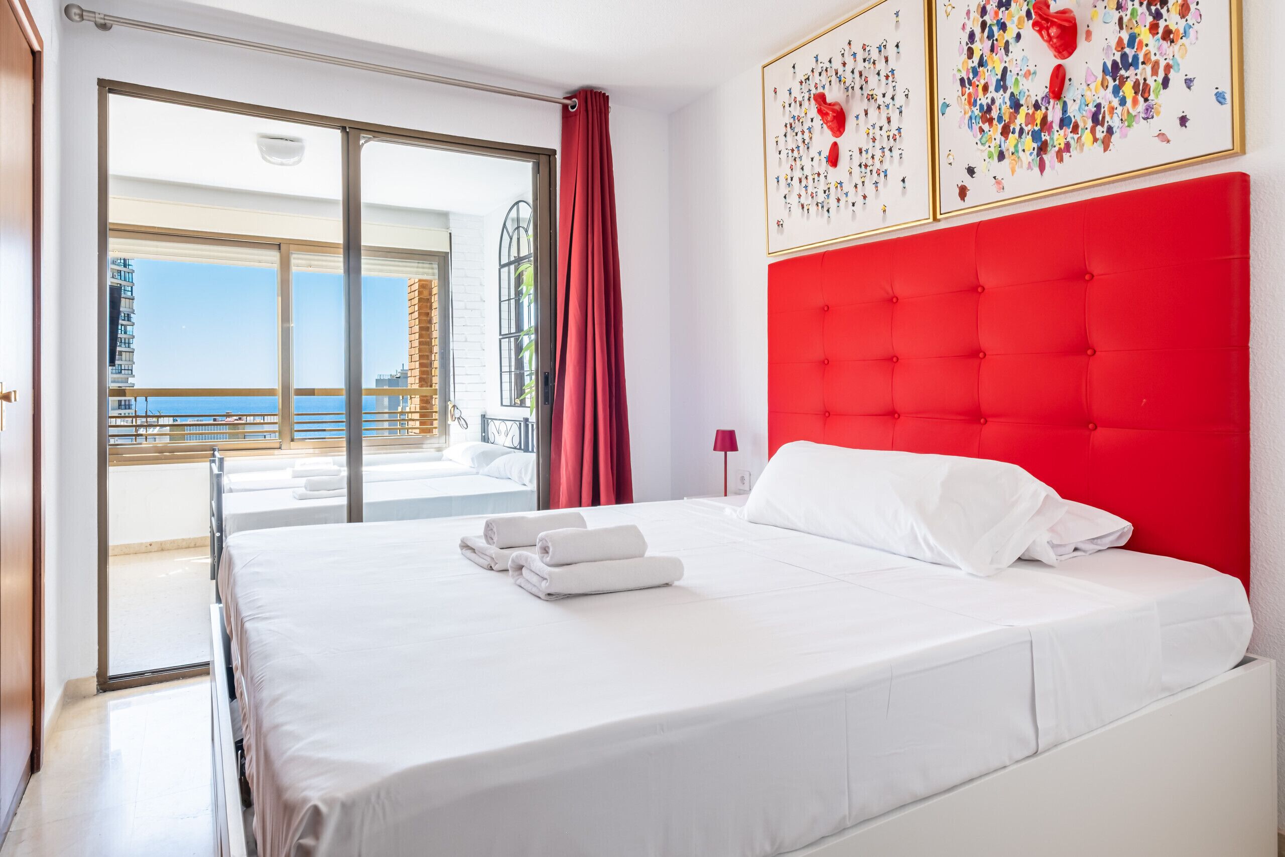 A hotel room with a bed, a red headboard, a balcony with a view, and artwork on the wall.