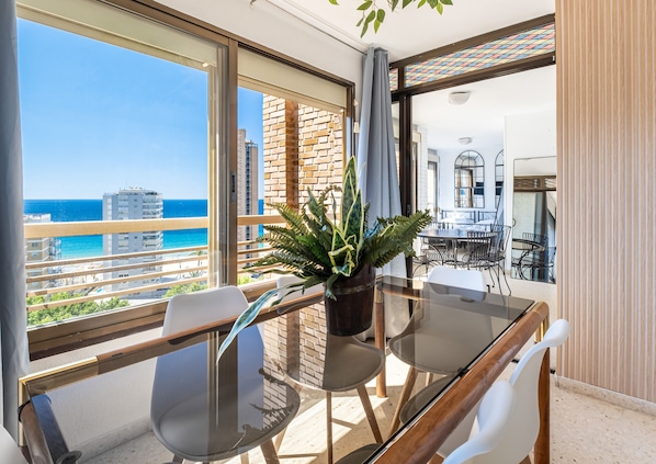 A balcony with a glass table, chairs, and a potted plant, overlooking a cityscape and the sea. - Las Rocas 14-A Apartment Levante Beach (Benidorm)