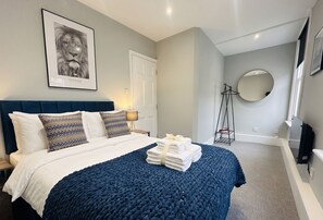 A bedroom with a bed, pillows, a blue blanket, a framed picture, a mirror, and a folded towel on the floor. - Vibrant Central One bedroom
67PRE01 (Brighton and Hove)