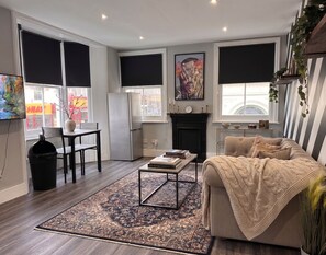 A modern living room with a sofa, a coffee table, a fireplace, and large windows with blinds. - Vibrant Central One bedroom
67PRE01 (Brighton and Hove)