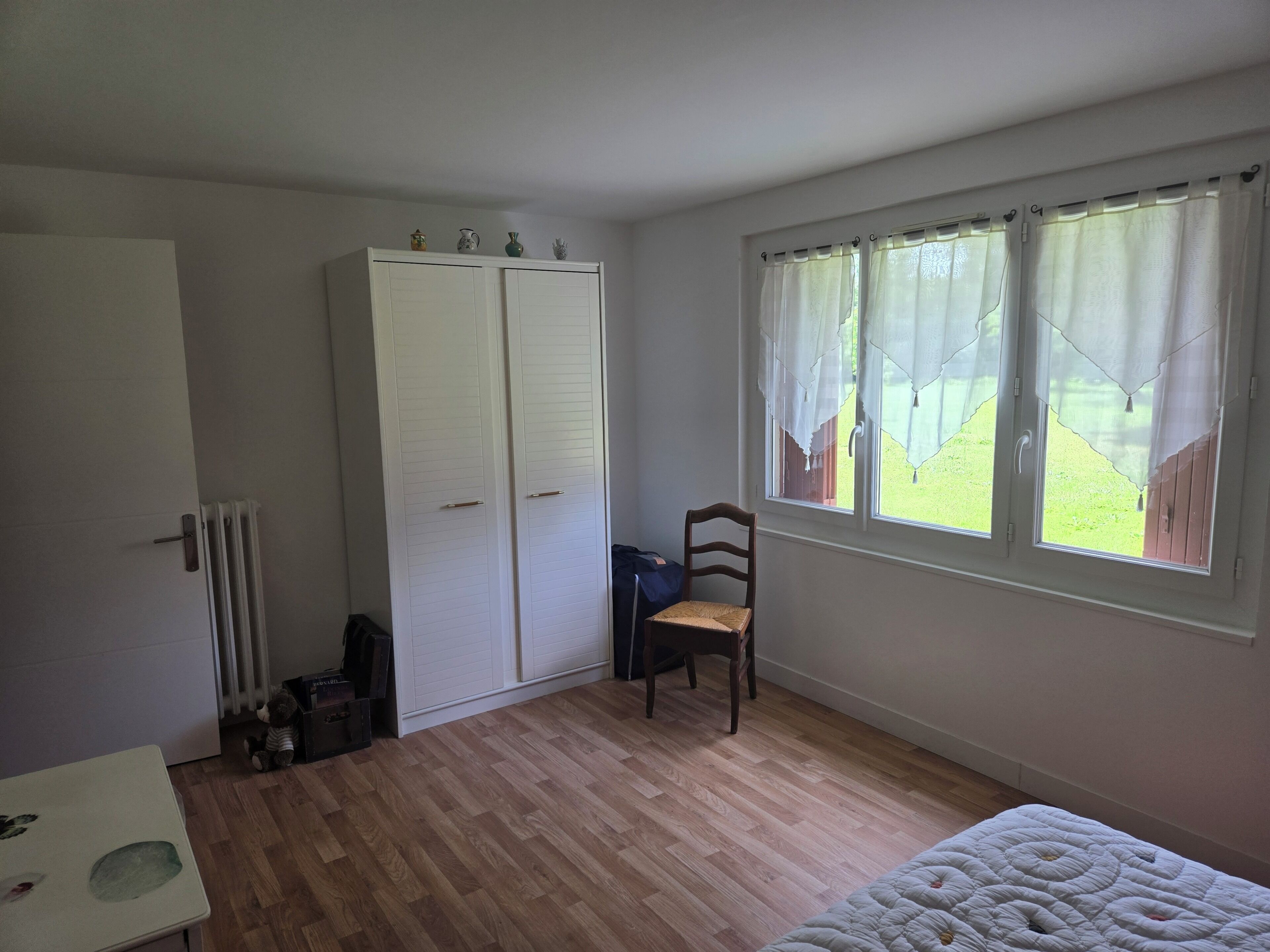 Une pièce avec un plancher en bois, un lit, une chaise, une armoire et une fenêtre donnant sur un jardin.