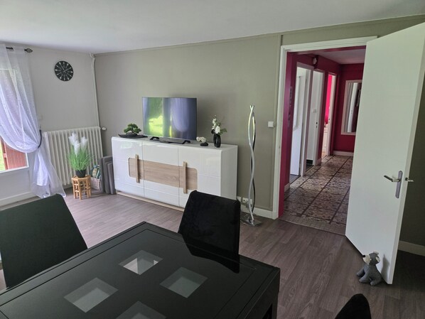 Una sala de estar con una mesa de comedor, un televisor en un mueble bajo, una planta en maceta y un pasillo que lleva a otra habitación.