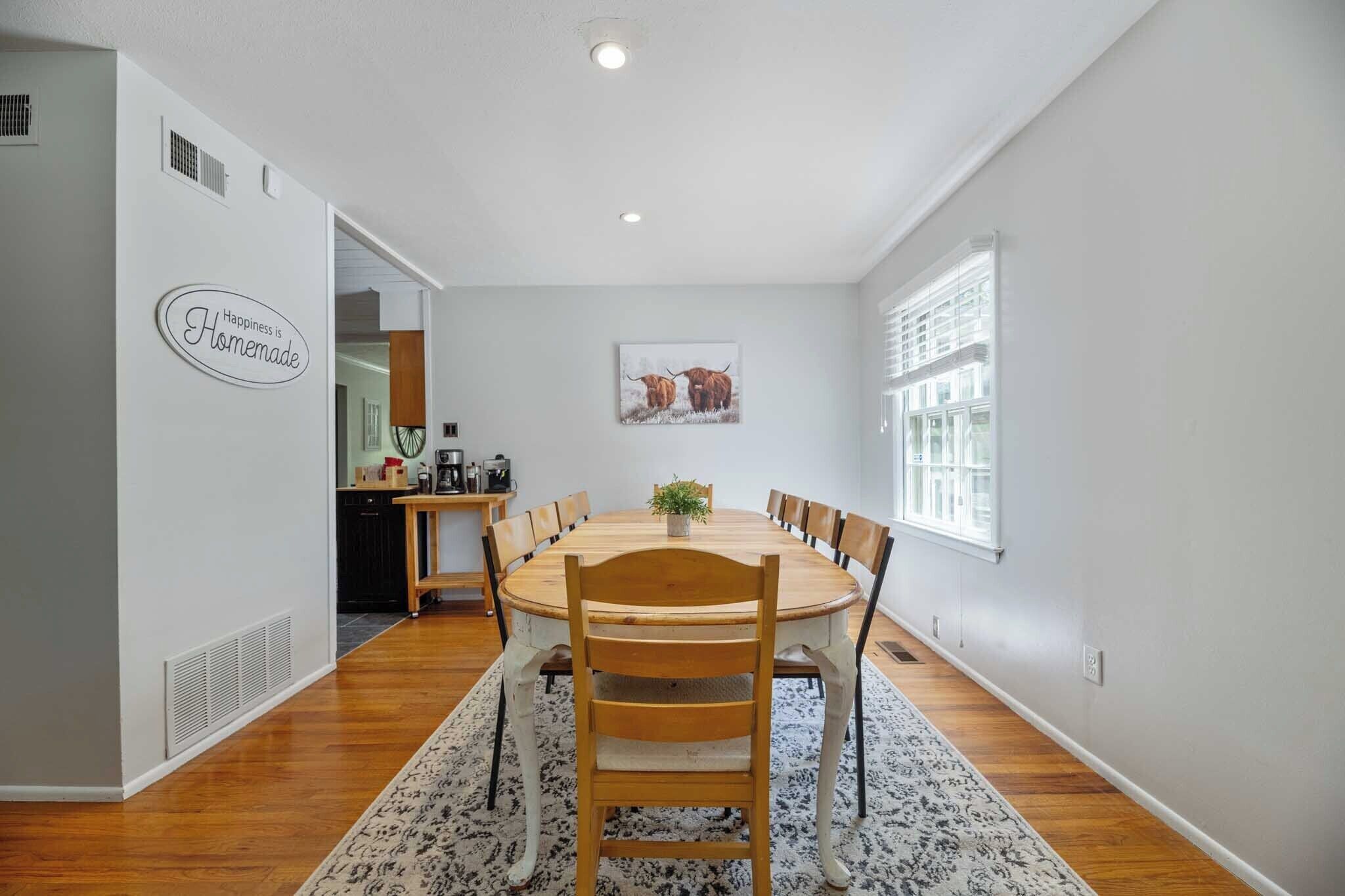 A dining area with a round table, chairs, a rug, a kitchen in the background, and a wall sign.