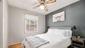 A bedroom with a bed, a ceiling fan, a window with blinds, a bedside table with a lamp, and a wall-mounted picture. - The Park House I Family Retreat Near Fort Benning & Auburn (Columbus)