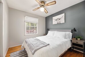 Una camera da letto con un letto, una ventola a soffitto, una finestra con persiane, un comodino con una lampada e un quadro appeso al muro.