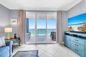 A room with a balcony, a sofa, a TV stand, and a painting of a beach. - Twin Palms 801 (Panama City Beach)