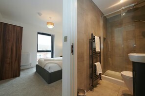 A modern bedroom with a large window, a bed, a wooden wardrobe, and a bathroom with a glass shower enclosure. -  2-Bed, 2-Bath Apartment in the Heart of York City Centre with secure parking (York)