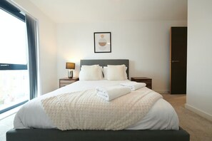 Een hotelkamer met een groot bed, een nachtkastje met lamp, een lijst met afbeelding aan de muur en uitzicht op de stad door het raam.