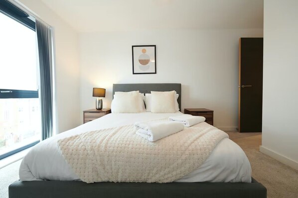 A hotel room with a large bed, a bedside table with a lamp, a framed picture on the wall, and a view of the city through the window. -  2-Bed, 2-Bath Apartment in the Heart of York City Centre with secure parking (York)