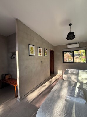 Ein Schlafzimmer mit einem Bett, einer Holzbank, einer Wand-Klimaanlage und einem Fenster mit Blick auf Grünflächen.