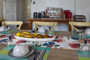 Un coin repas avec une table dressée pour quatre personnes, un panier de fruits et divers ustensiles de cuisine disposés sur le comptoir.
