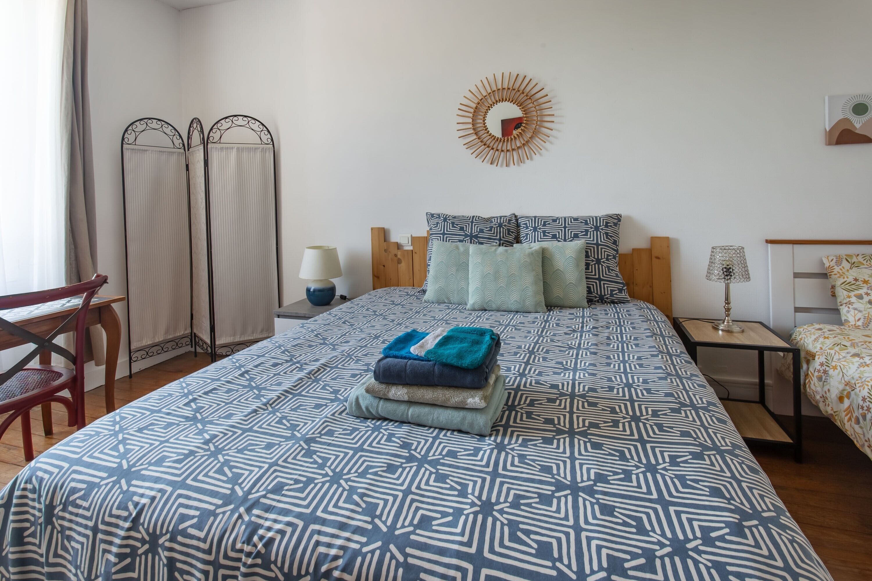 A neatly arranged bedroom with a patterned bedspread, a bedside table with a lamp, a wall-mounted mirror, and a window with curtains.