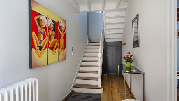 A hallway with a staircase, a radiator, a painting, a vase with flowers, and a mirror on the wall.