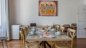 Zona pranzo con un tavolo lungo apparecchiato per un pasto, circondato da sedie in vimini, un quadro al muro e una porta bianca sullo sfondo.