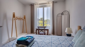 A bedroom with a bed, a wooden hanger rack, a table, and a window with curtains.