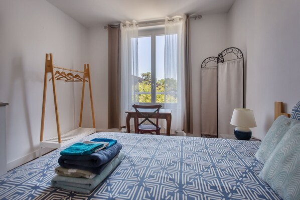 Un dormitorio con cama, perchero de madera, una mesa y una ventana con cortinas.