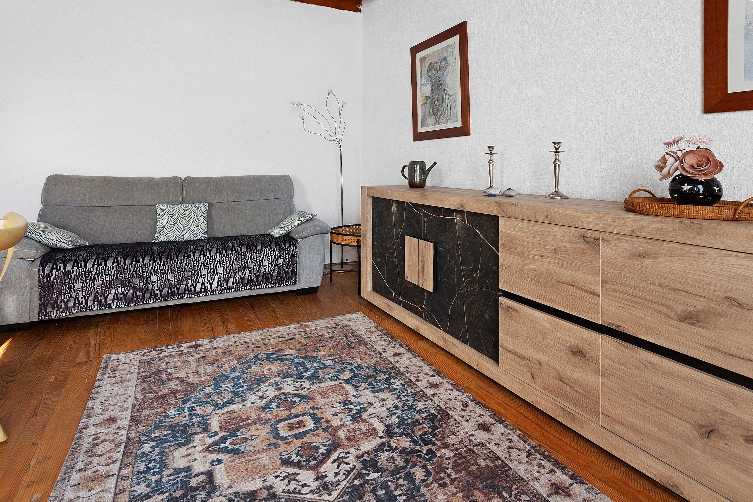 Un salon avec un canapé gris, des coussins à motifs, un meuble en bois, un tapis et un tableau encadré accroché au mur.