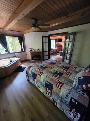 A bedroom with a wooden ceiling, a quilted bedspread, a wooden floor, a window with curtains, and a door leading to another room. - Eagles Nest Cabin (New Straitsville)