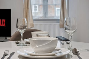 A hotel room with a dining table set with white plates, bowls, cutlery, and two empty wine glasses. - Modern & Historic Mill Flat with Stream Views (Surrey)