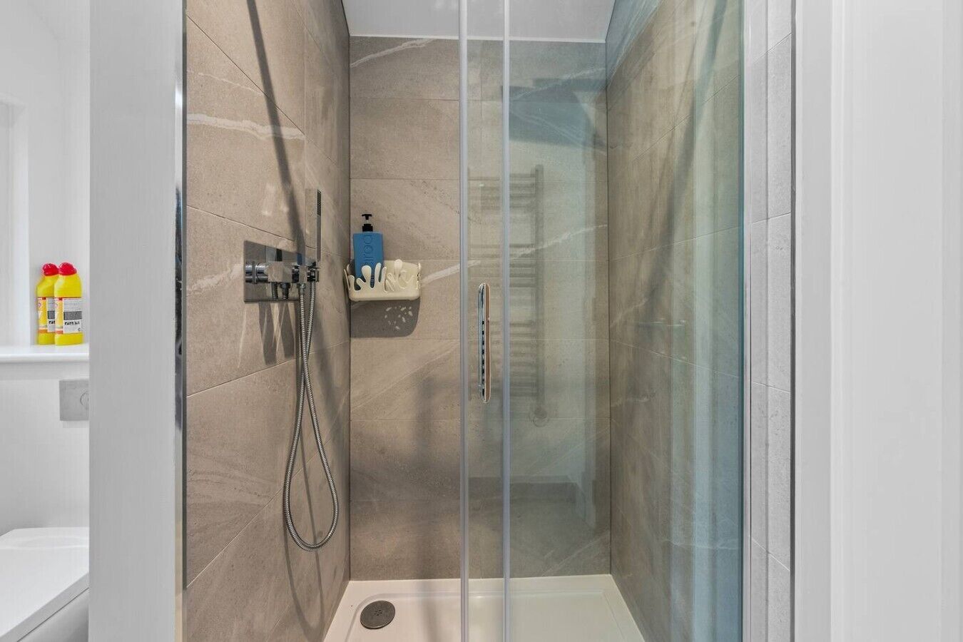 Une douche fermée par des parois en verre, avec des carreaux de couleur beige, une douchette à main et un distributeur de savon.