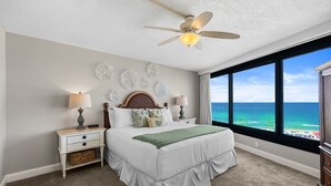 Un dormitorio con cama, mesitas de noche, ventilador de techo, decoración en la pared y un amplio ventanal con vista al océano.