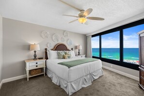 Ein Schlafzimmer mit einem Bett, Nachttischen, einem Deckenventilator, Wandkunst und einem großen Fenster mit Meerblick.