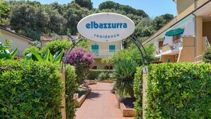 Ein Weg führt zu einem Gebäude mit der Aufschrift 'elbazzurra residence', mit einem Schild, umgeben von Grünpflanzen und blauem Himmel.