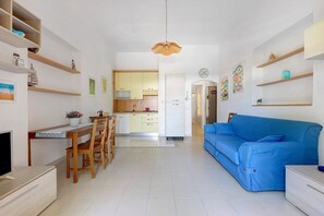 A modern kitchen with white cabinets, a dining table with chairs, a blue sofa, and a TV on a wooden stand.