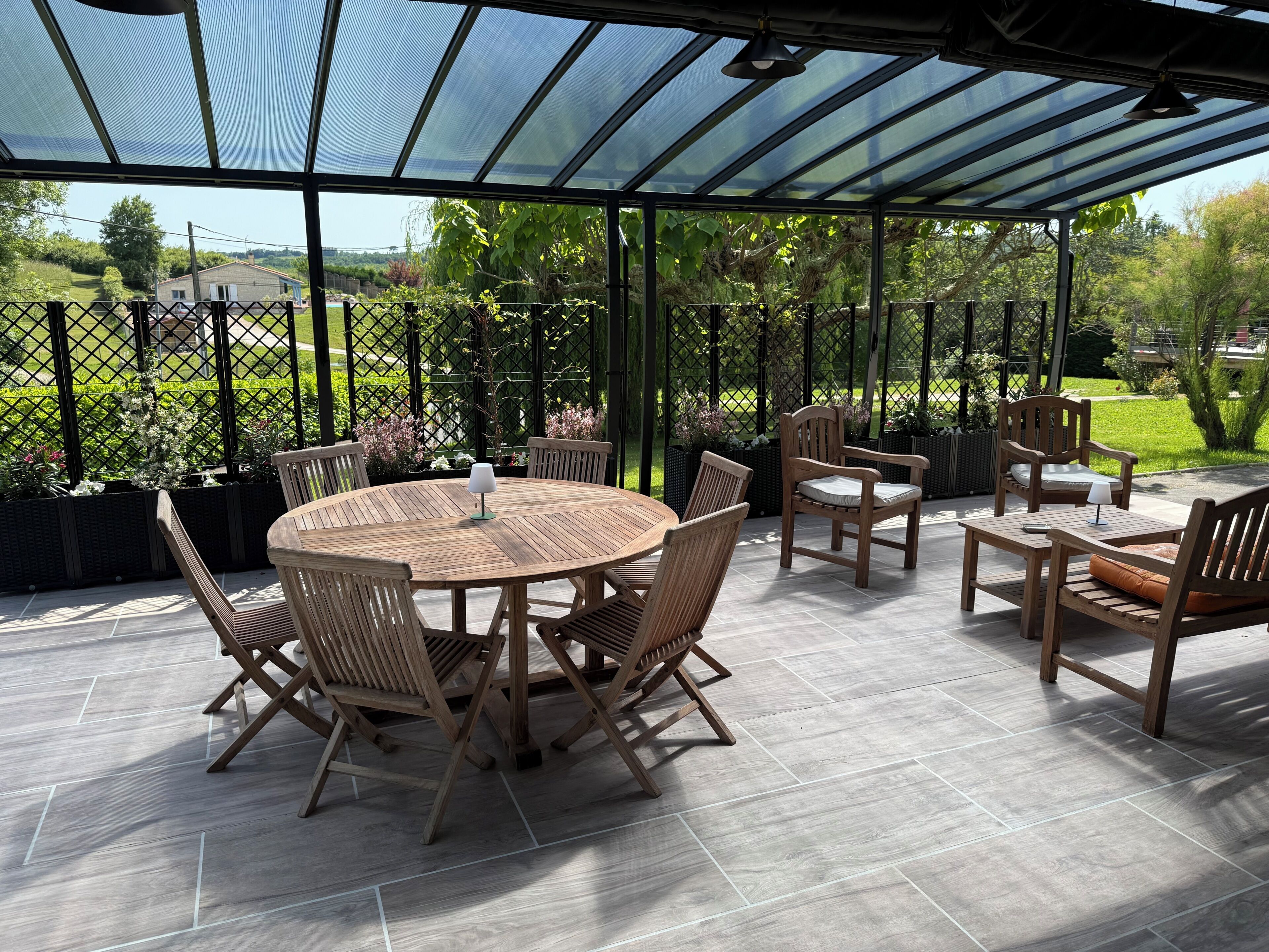 Uma área de varanda coberta com uma mesa redonda de madeira e cadeiras, uma mesinha com um abajur e um banco.