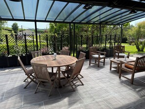 Een overdekt terras met een ronde houten tafel en stoelen, een kleine tafel met een lamp, en een bank.