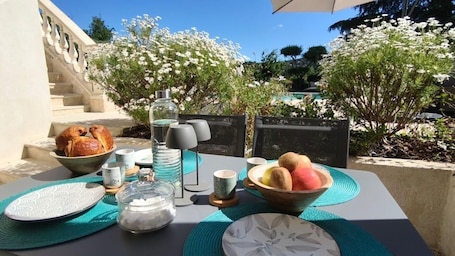 An outdoor dining setup with a table, plates, cups, a bowl of fruit, and a bottle.. Large studio in villa with swimming pool, jacuzzi and boules pitch.