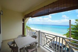 Ein Balkon mit Tisch und Stühlen, mit Blick auf einen Strand mit klarem, blauem Wasser und sandigem Ufer.