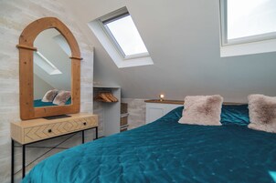 Un dormitorio con techo inclinado, un espejo grande, una cama con colcha color turquesa y un tragaluz.