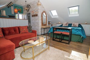 A cozy room with a red sofa, a glass coffee table, a bed with teal bedding, and a loft-style ceiling with skylights. - Garden View Apartment (Kingsbridge)