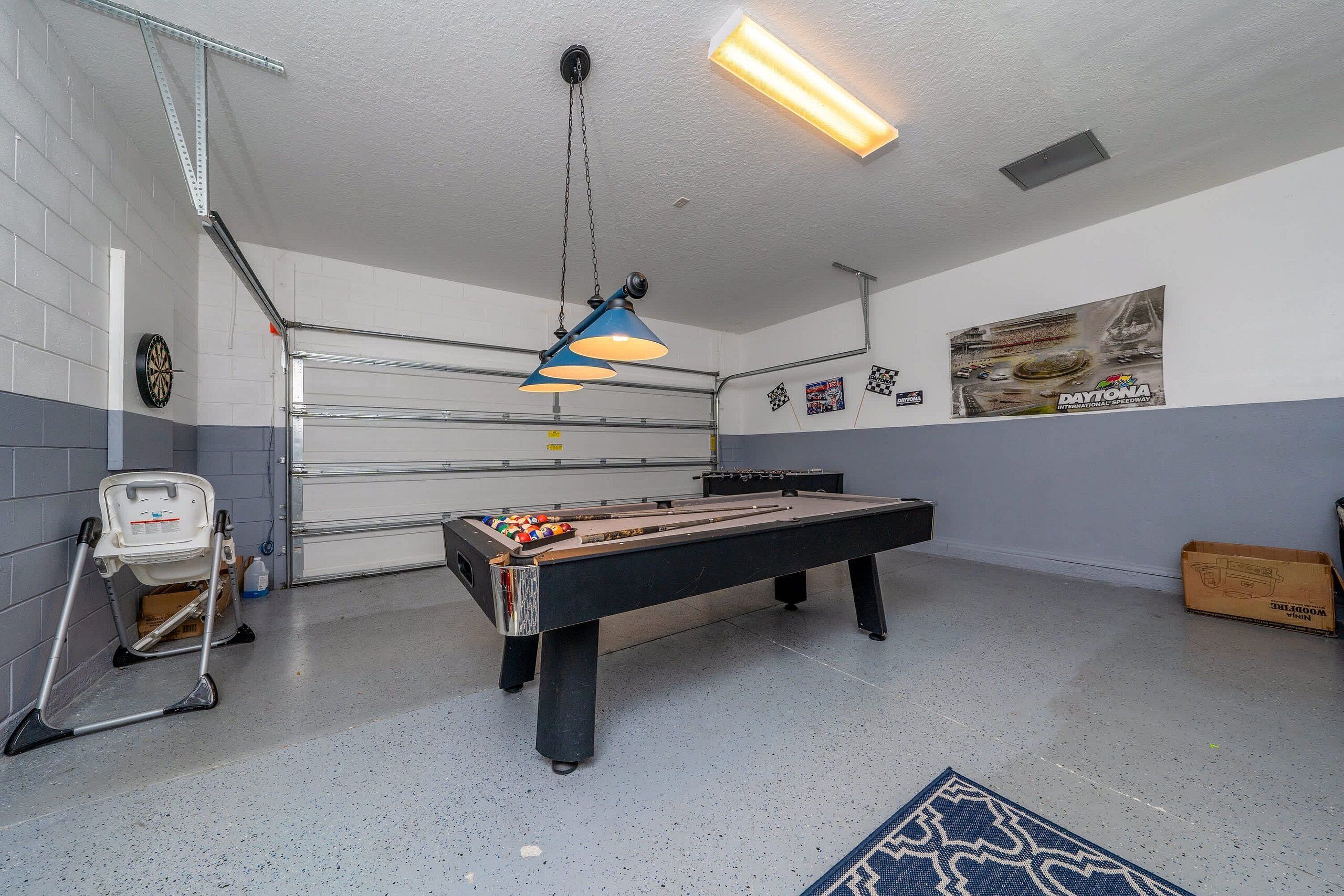 A pool table with a blue overhead light, a dartboard, and a poster on the wall.
