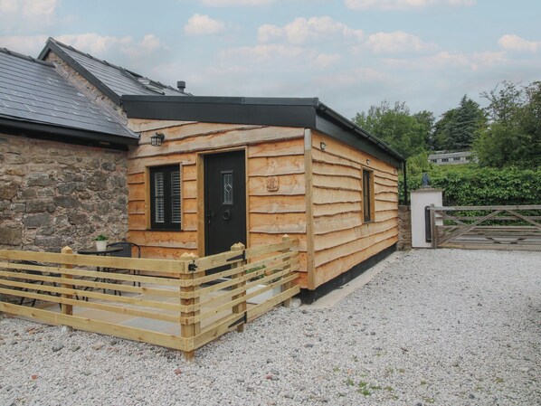 A wooden cabin with a stone wall, a black door, and a gravel driveway. - The Dinky Dairy (Oswestry)