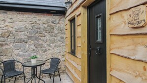A wooden building with a stone wall, a black door, and a sign that reads 'Dinky Dairy.' There is a small table with two chairs outside. - The Dinky Dairy (Oswestry)