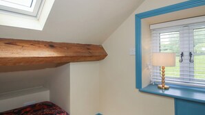 A room with a sloped ceiling, a window with blinds, a bedside table with a lamp, and a patterned bedspread. - The Dinky Dairy (Oswestry)