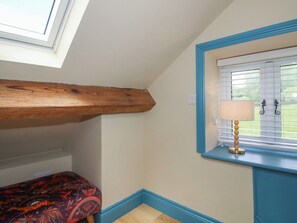 A room with a sloped ceiling, a window with blinds, a bedside table with a lamp, and a patterned bedspread. - The Dinky Dairy (Oswestry)
