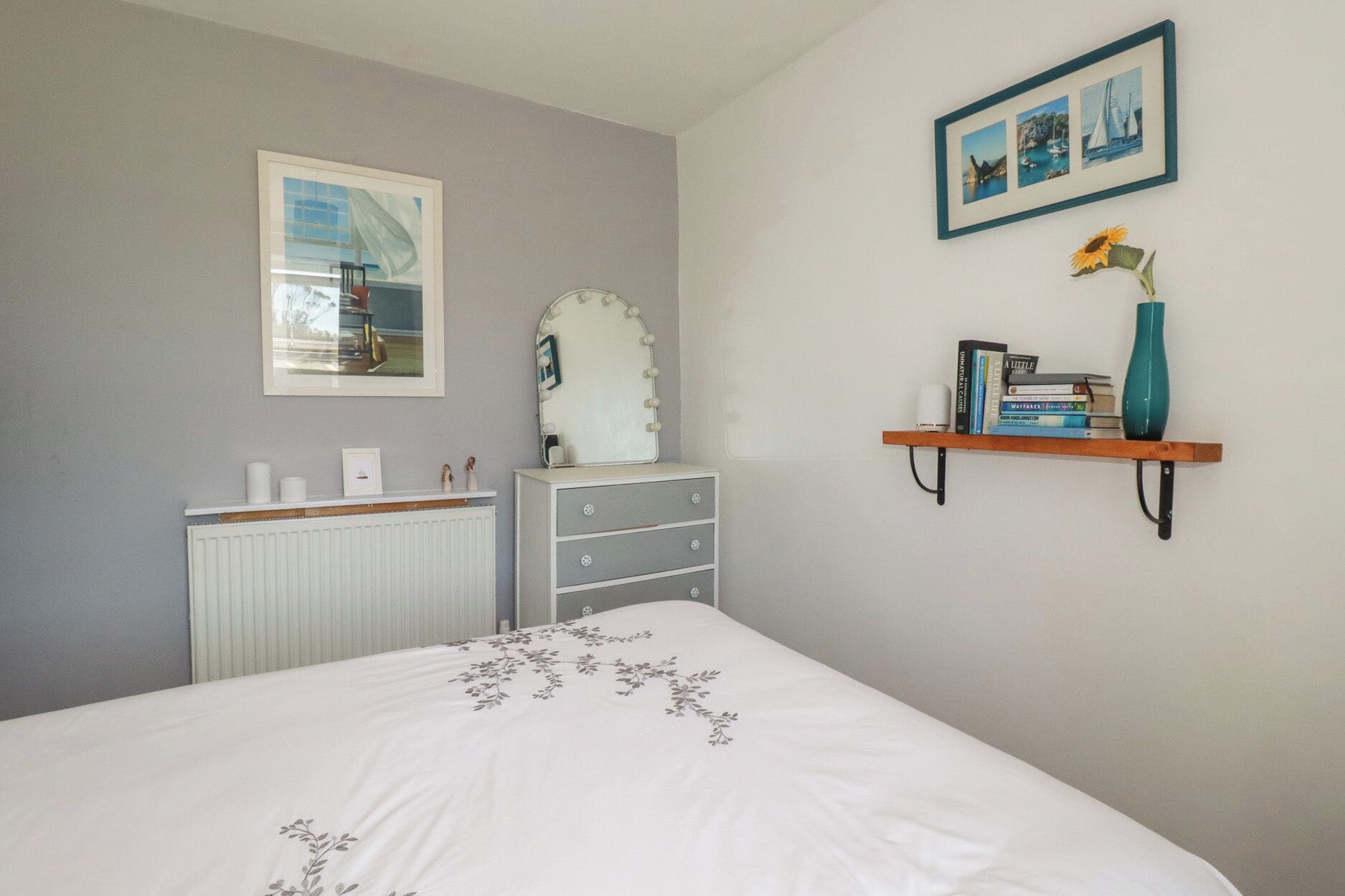 Un dormitorio con cama, cómoda, tocador, radiador, cuadros enmarcados y una estantería con libros y un florero.