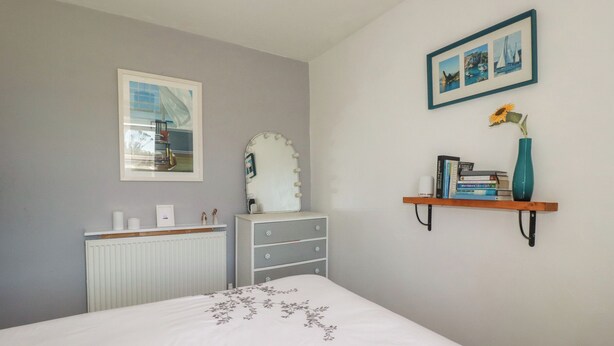 A bedroom with a bed, dresser, vanity, radiator, framed pictures, and a shelf with books and a vase. - Tin Lantern Cottage (Redruth)