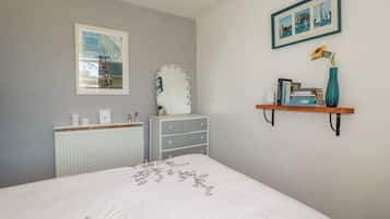 Un dormitorio con cama, cómoda, tocador, radiador, cuadros enmarcados y una estantería con libros y un florero.