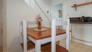 A dining area with a wooden table and chairs, a vase with flowers, a staircase, and a kitchen area with a shelf. - Tin Lantern Cottage (Redruth)